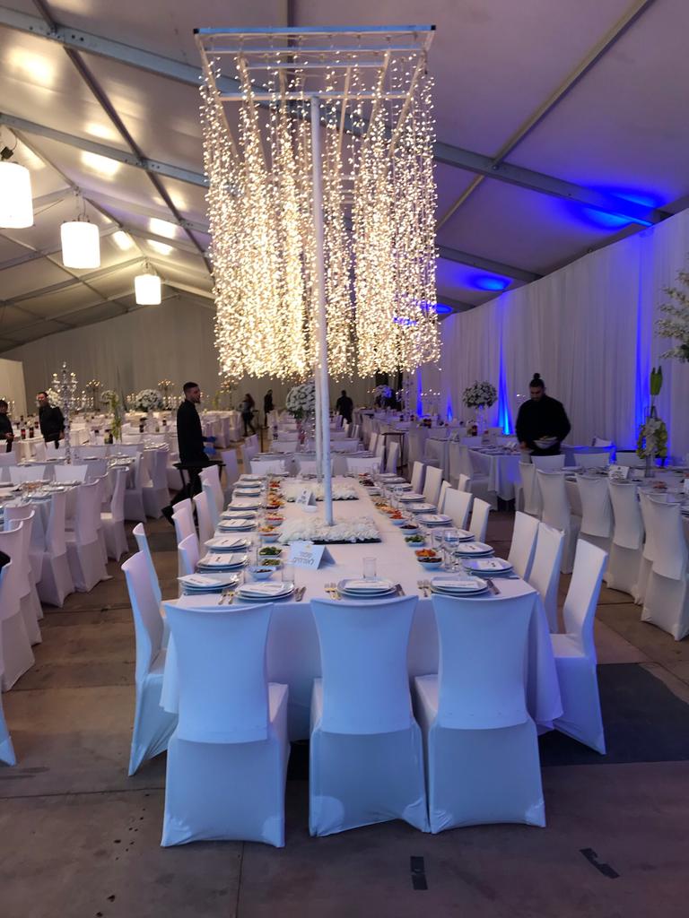 שולחנות ערוכים לאירוע עם מפות וכסאות לבנים וקישוטים תלויים של נורות קטנות.